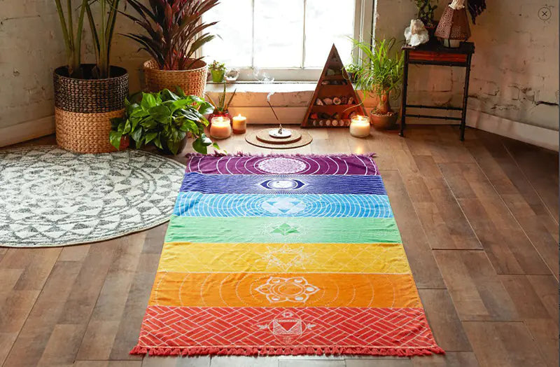 Colorful chakra tapestry on a wooden floor with plants and candles in the background. From ghg wellness shop.