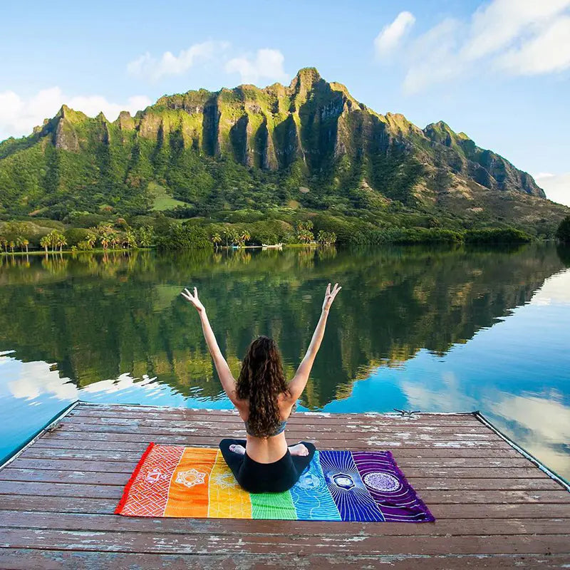 Person meditating on a dock with colorful towels, surrounded by a mountainous landscape. From ghg wellness shop.